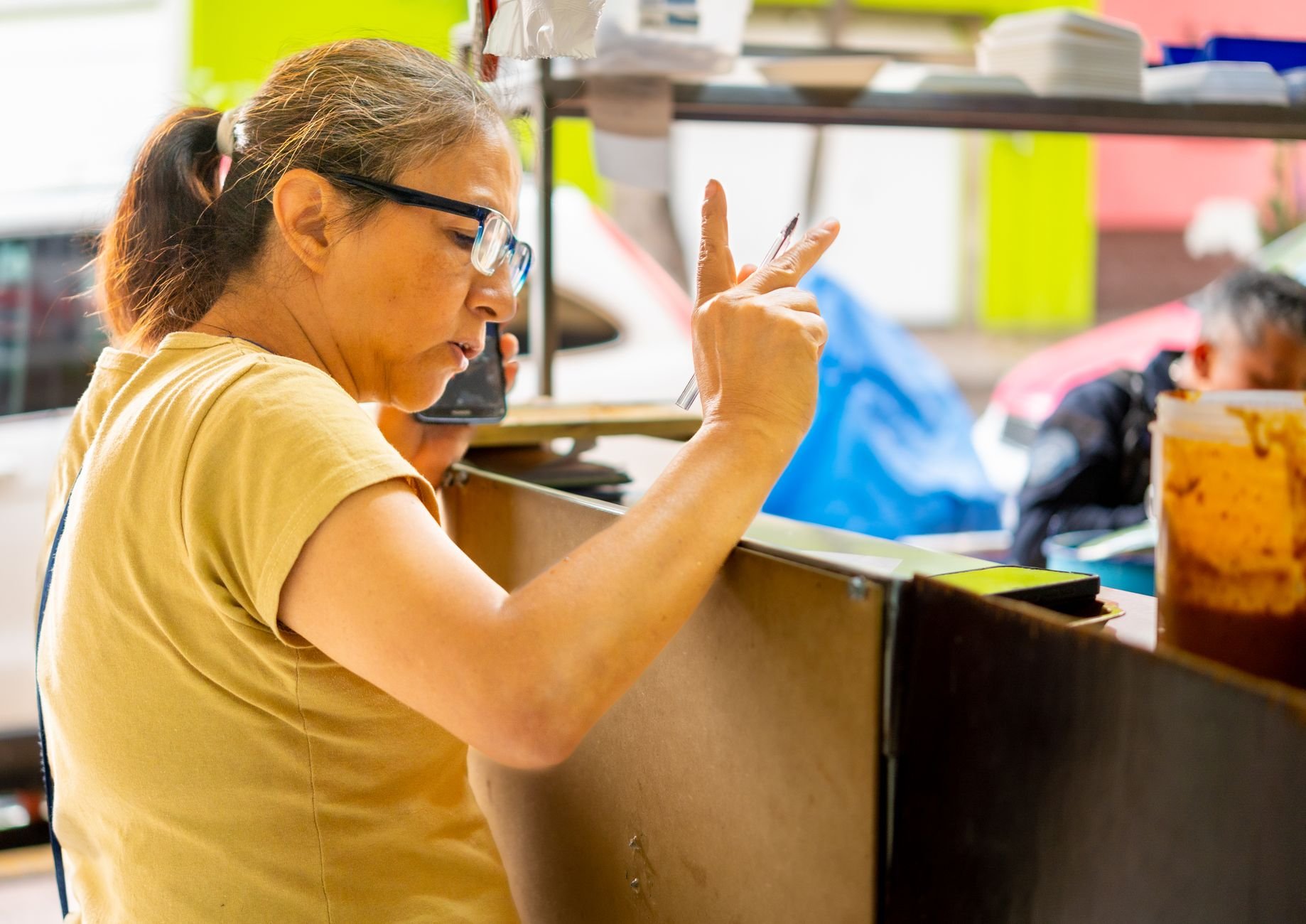 A female vendor takes an order using a smartphone at a lively outdoor food stall